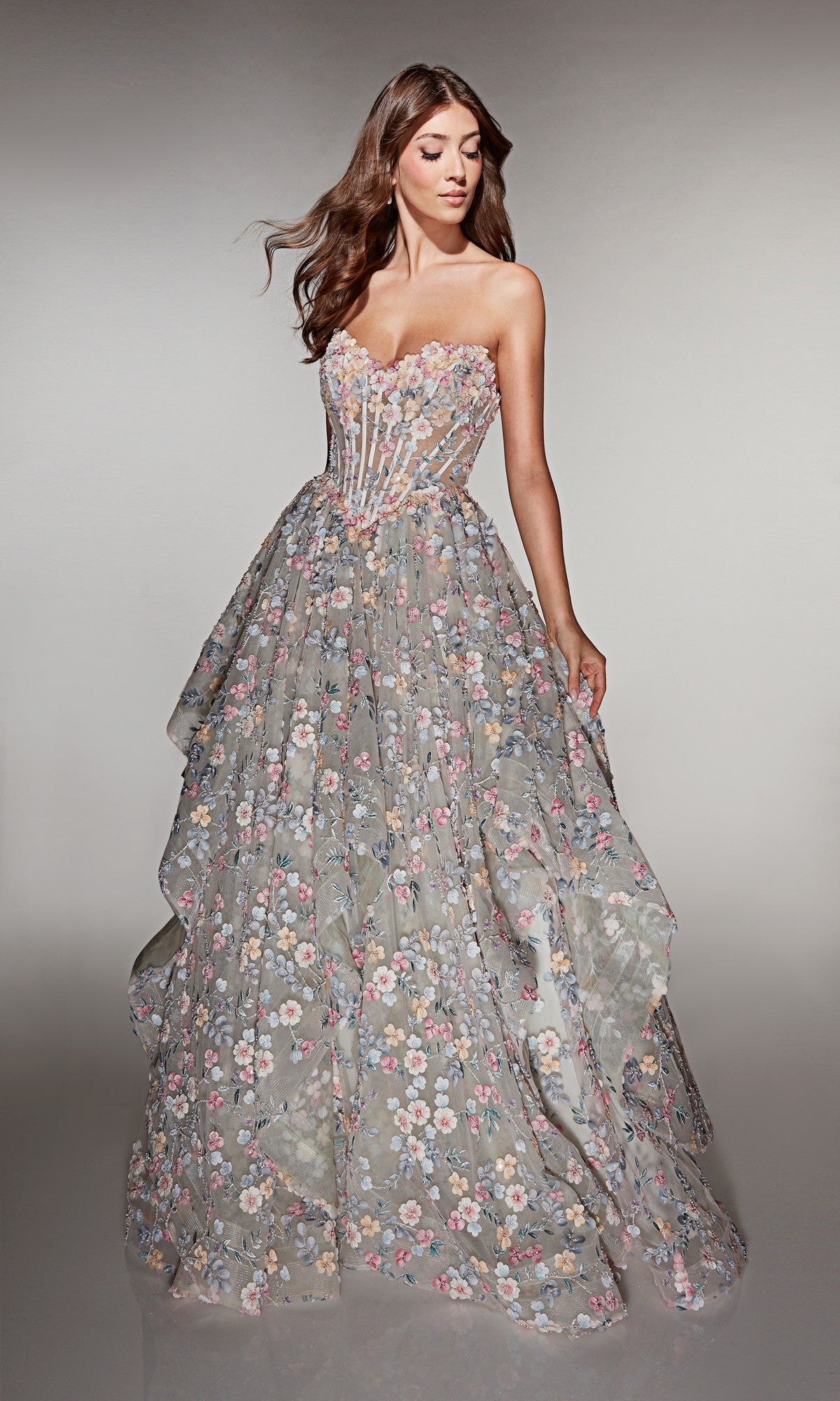A woman stands against a neutral background wearing the Alyce 62048, a long prom dress featuring a strapless corset bodice and a full floral-patterned skirt.